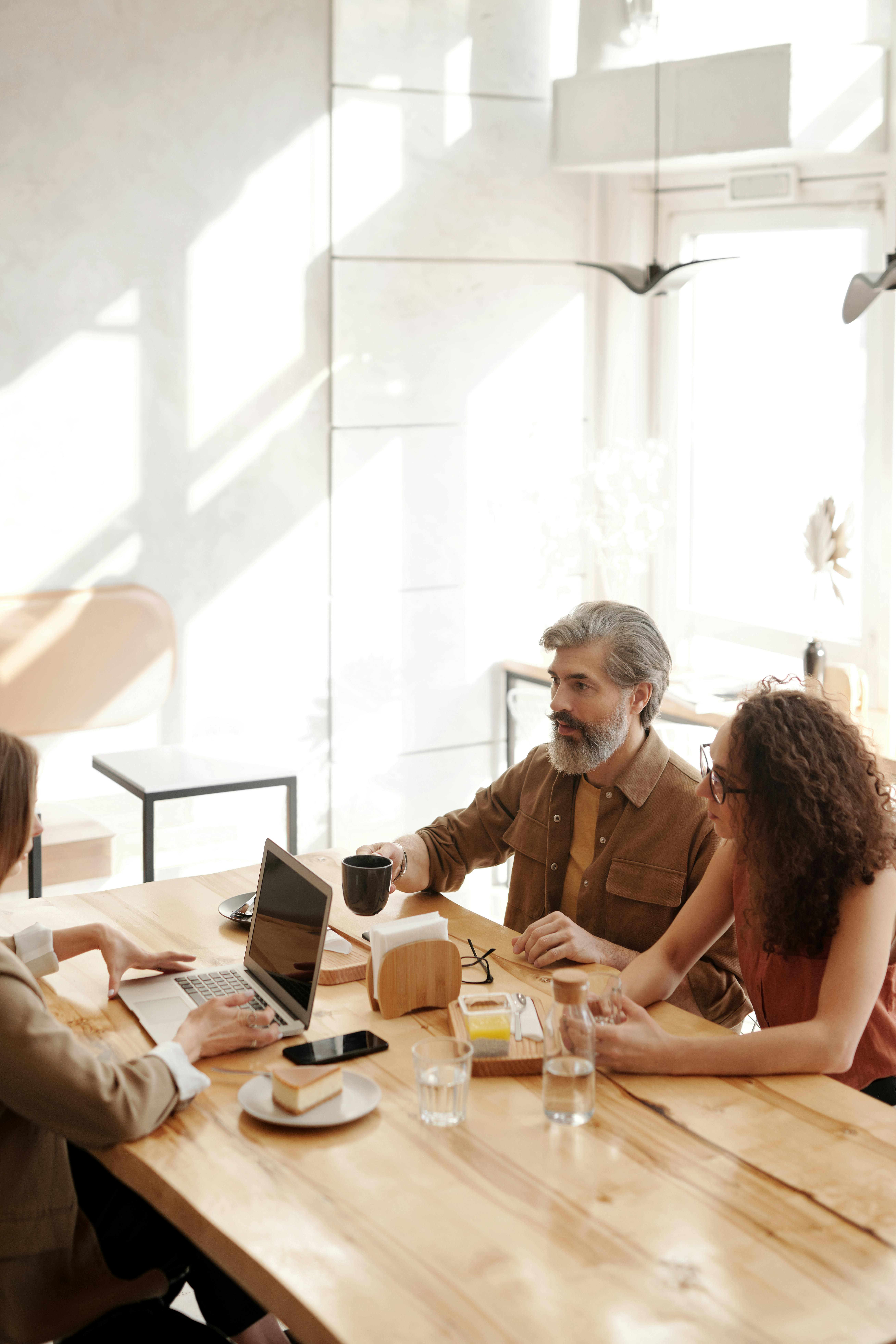 Three colleagues in a coffee shop discussing work at a table with laptops and drinks.